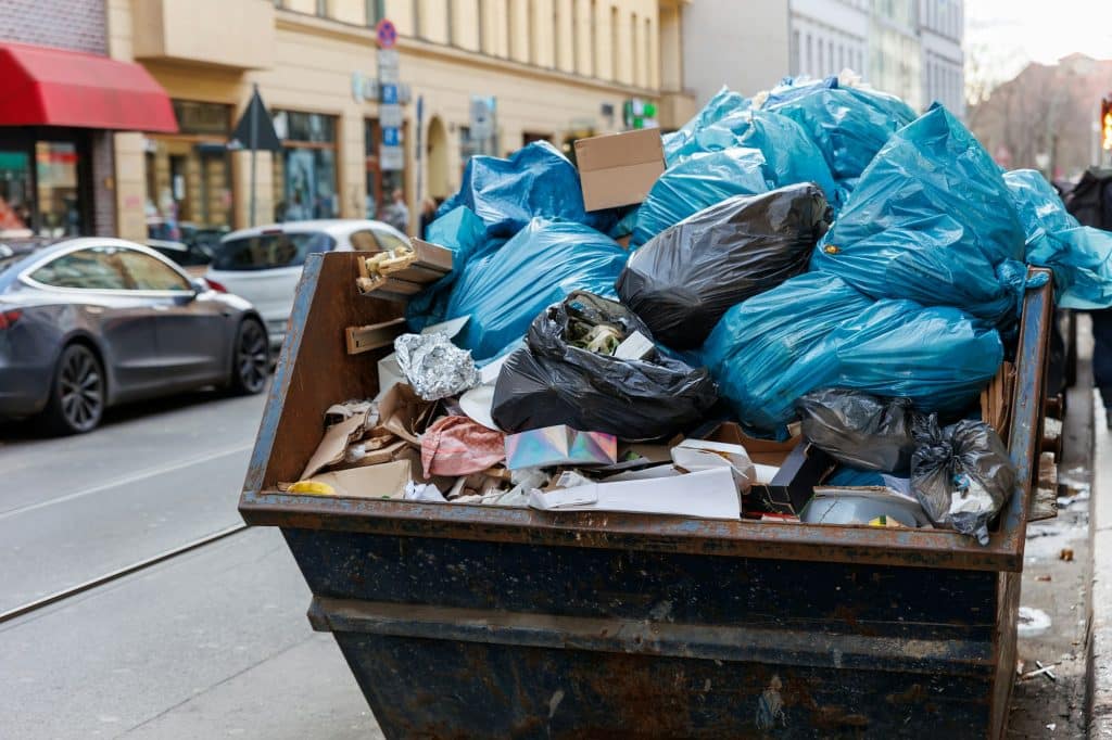 Big metal overloaded dumpster skip container full of plastic trash litter bags near residential